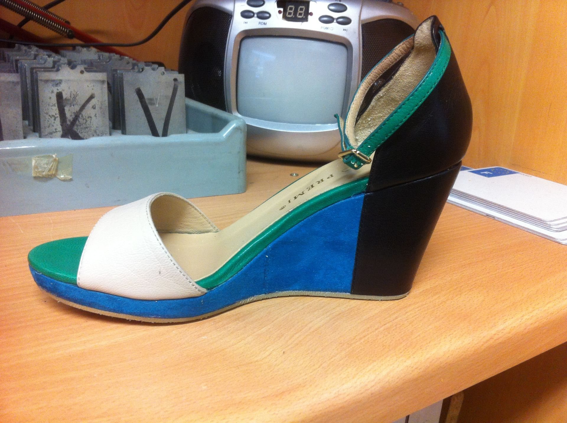 Sandale à talon compensé en cuir beige, vert, bleu et noir posée sur une étagère en bois, devant un petit poste radio avec écran.